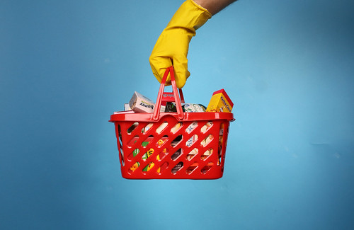 'Follow Shopping basket and hand in a rubber glove isolated on blue background. Safe shopping concept.' by Jernej Furman